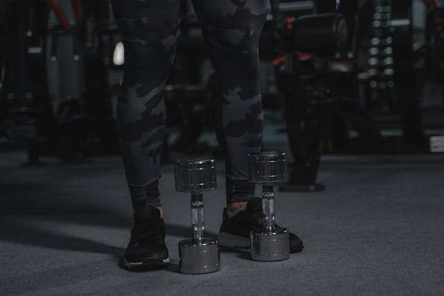 A person standing beside a pair of dumbbells on grey gym flooring.