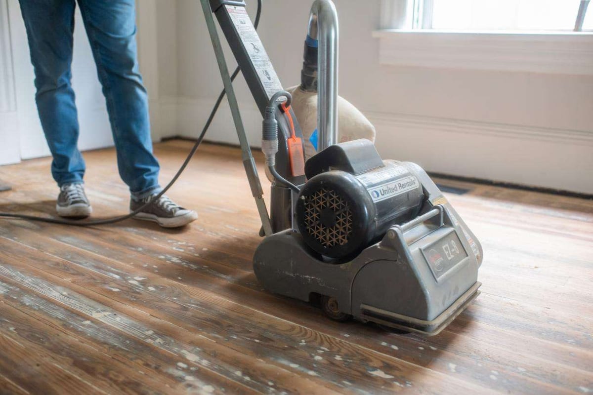 A drum sander being run along an old hardwood floor during a refinishing job, with sanding dust on the boards.