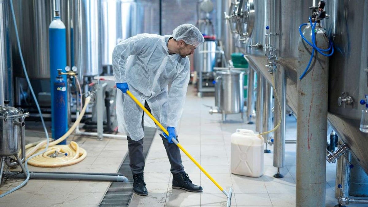 A worker in protective clothing mopping the floor of an industrial brewery.