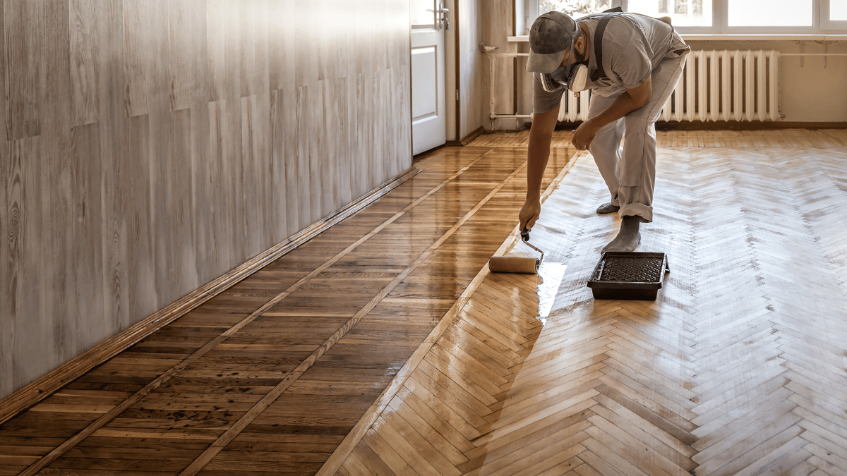 A worker applying finish with a roller to a herringbone wood floor.