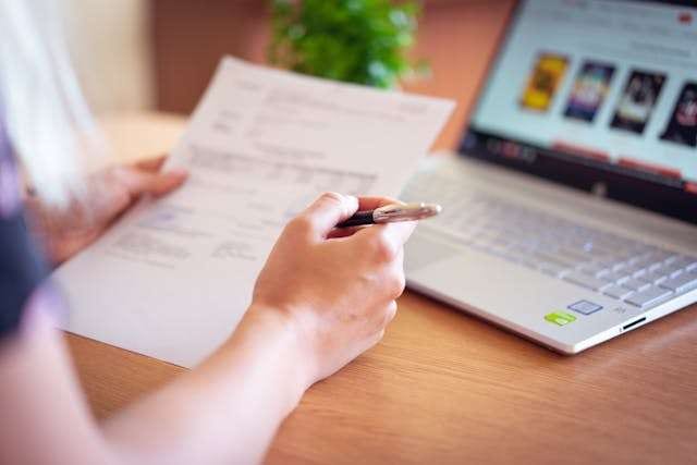 A person reviewing a printed document with a pen beside a laptop.