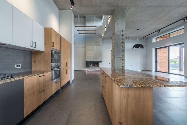 A long kitchen with oak cabinetry, a granite island, concrete columns and dark large-format tile.