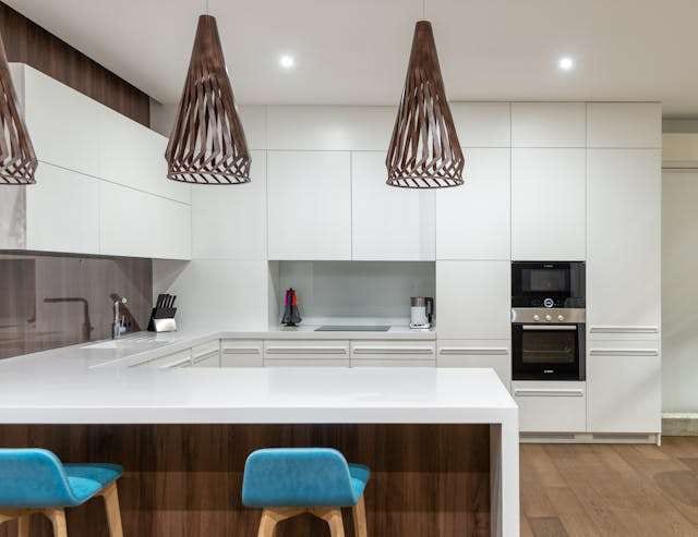 A white kitchen with a wood-fronted island, blue stools and wooden pendant lights.