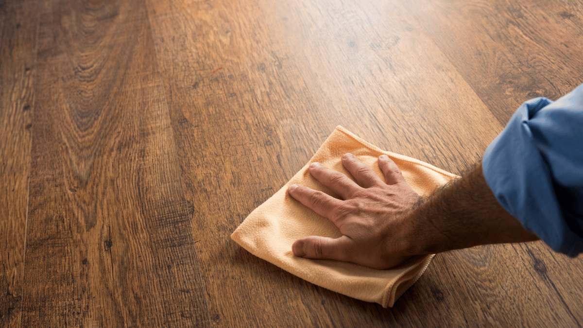 A hand wiping a dark brown wood-look plank floor with a soft yellow cloth.