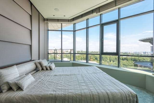 A bedroom with grey carpet and a wall of windows overlooking a city park.
