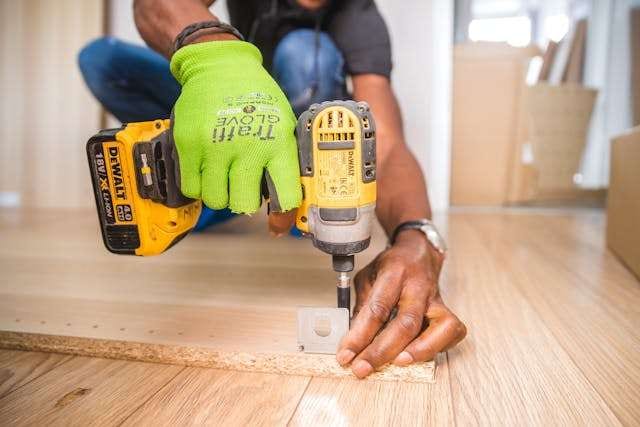 A gloved hand driving a screw with a cordless drill into a panel lying on a light wood floor.
