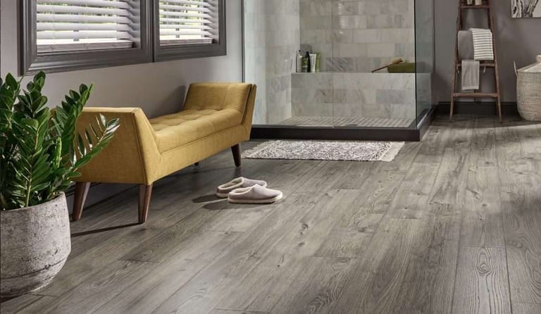Grey weathered wood-look plank flooring with a yellow bench, potted plant and slippers beside a tiled shower