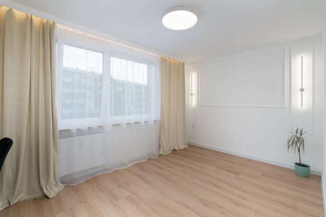 An empty room with light oak-look laminate flooring and cream curtains.