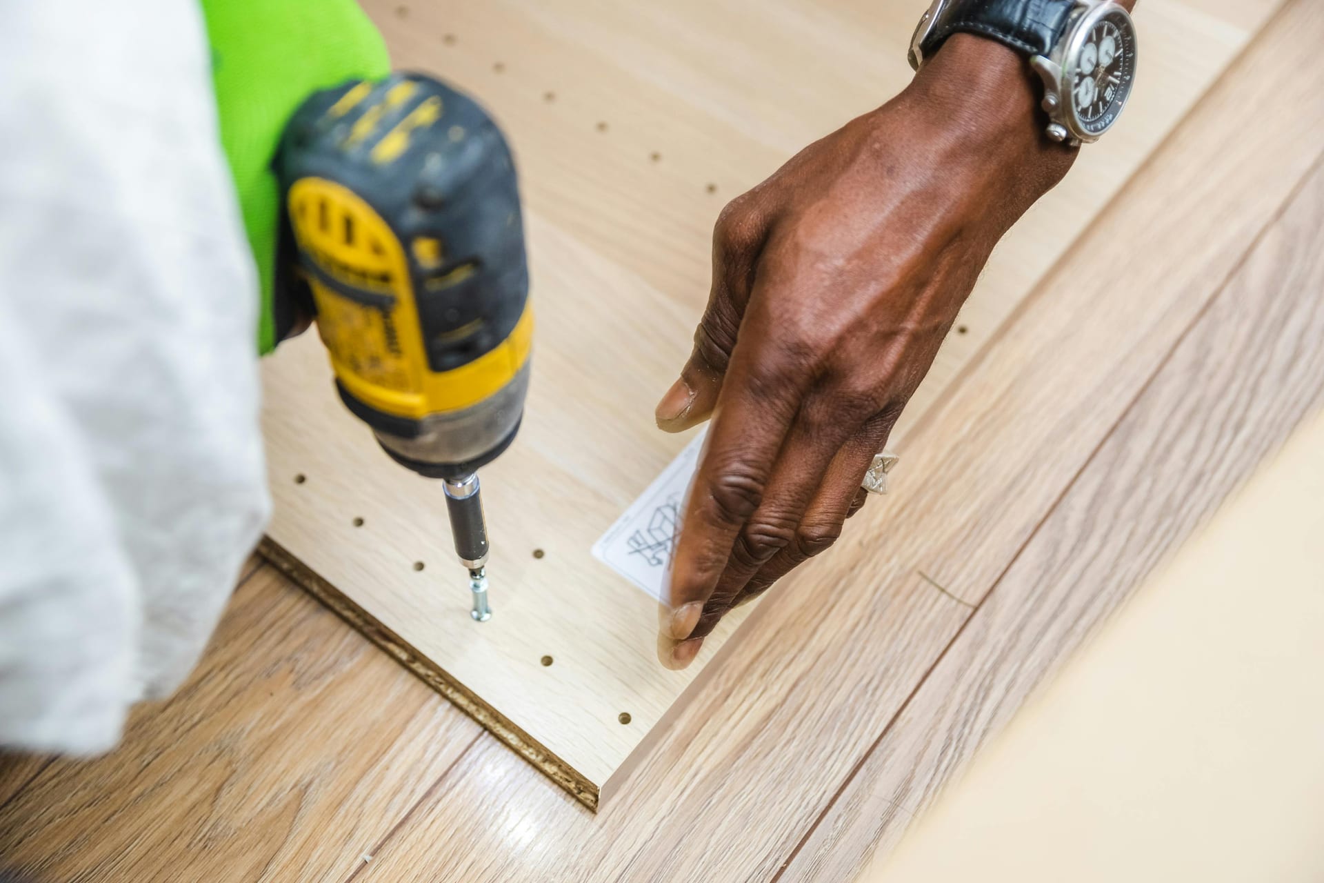 A gloved hand drilling a screw into a perforated wood panel.