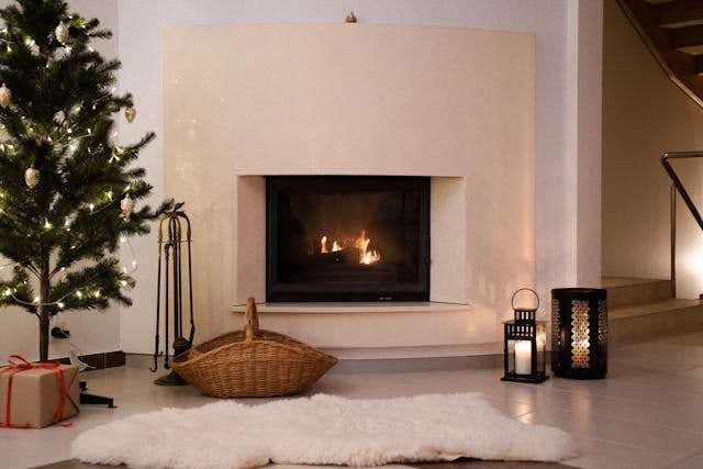 A cream fireplace surround with a lit fire, a Christmas tree and a sheepskin rug.