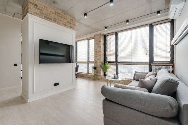 A loft living room with pale grey wood-look flooring, a grey sofa and a brick wall.