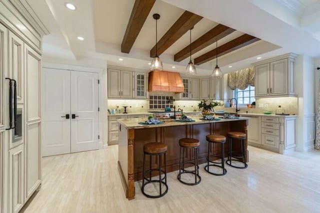 Kitchen with cream cabinetry, a dark wood island with leather stools, exposed timber ceiling beams and a pale travertine-look stone floor.