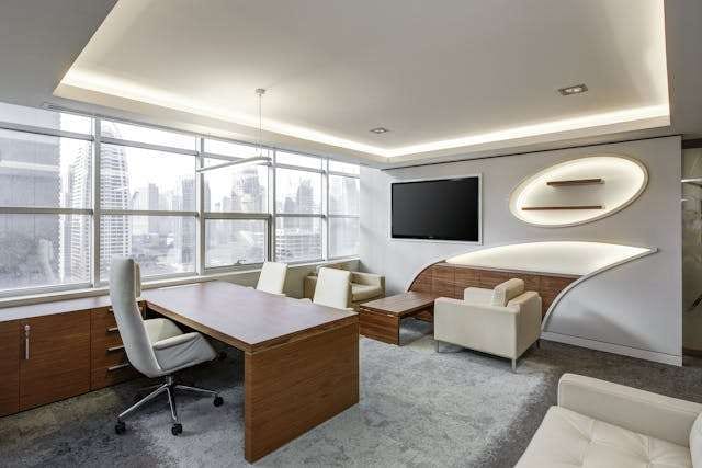 An executive office with grey carpet, a wooden desk and white armchairs.