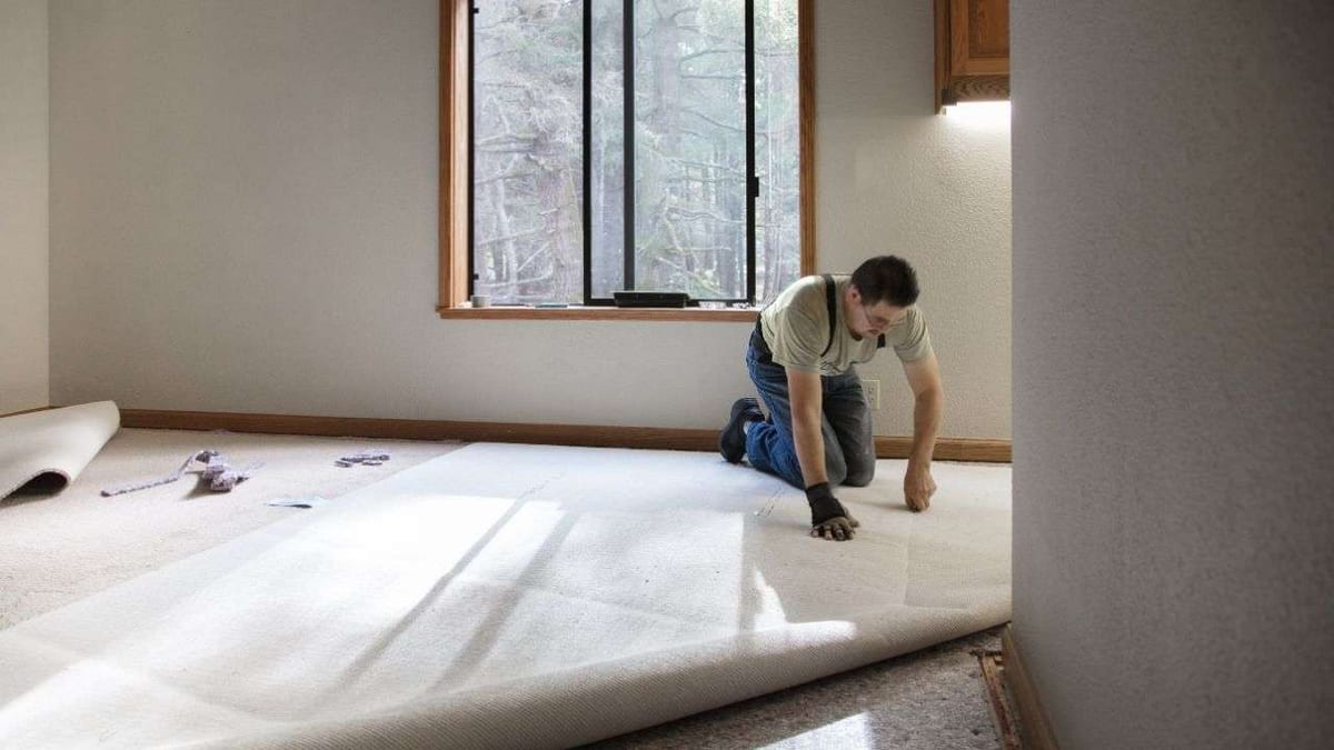 An installer kneeling to roll out new pale carpet in an empty room.