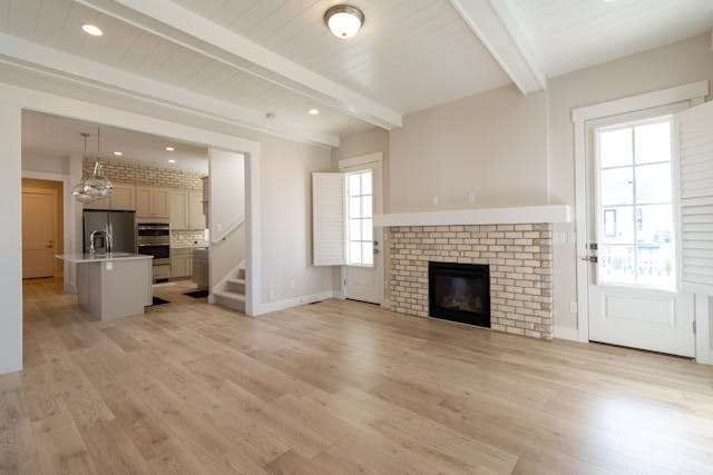 An empty living room with light wood-look flooring, a brick fireplace and a kitchen beyond.