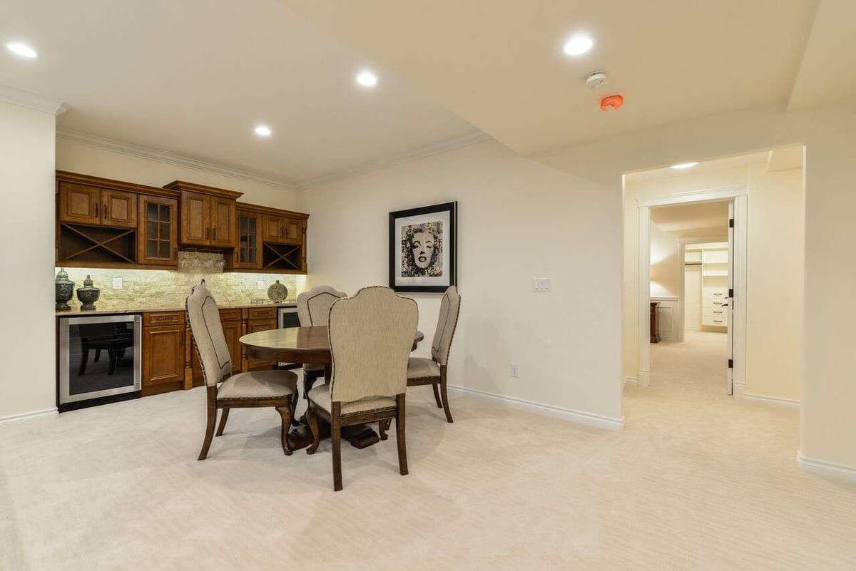 A basement games room with cream wall-to-wall carpet, a round table with upholstered chairs and a wet bar.