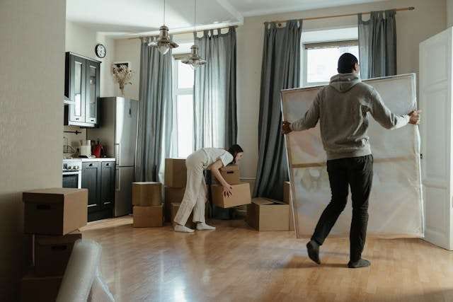 Two people carrying boxes and a framed picture across a light wood floor while moving.