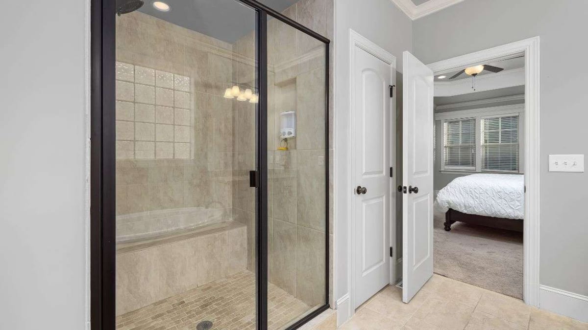A glass-enclosed shower with beige tile, opening onto a carpeted bedroom.