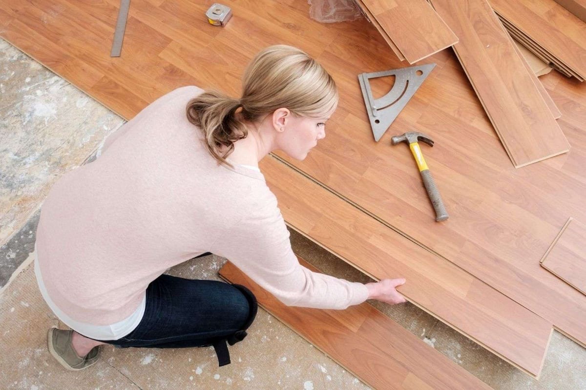 A woman kneeling to fit a laminate plank, with a hammer and a set square on the subfloor beside her.