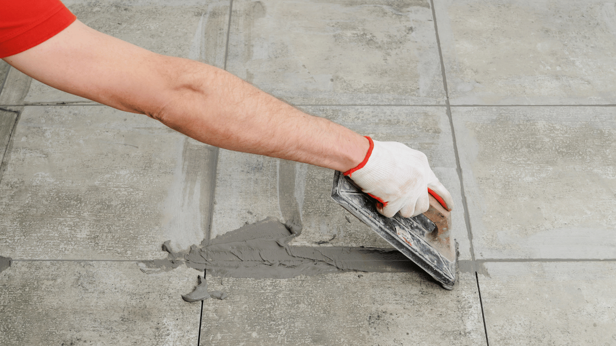 A gloved hand spreading grey grout across floor tile with a rubber float.