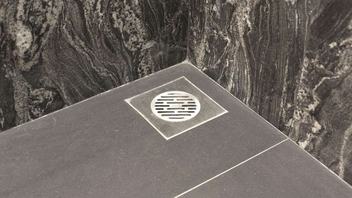 A grey stone shower floor with a square centre drain against a veined dark marble wall.